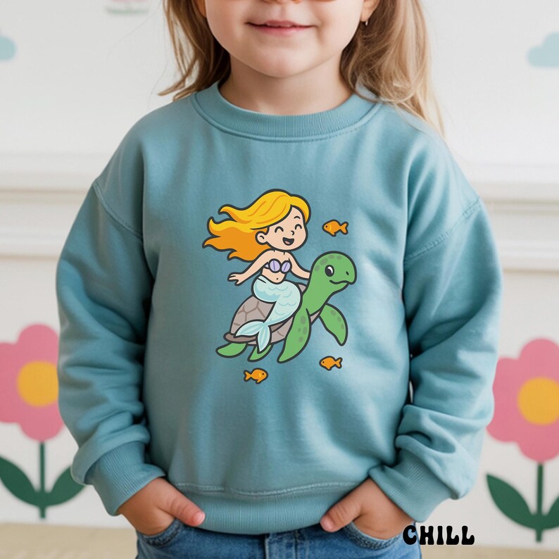 Puede incluir: Sudadera azul claro con una sirena de dibujos animados montada en una tortuga verde, rodeada de peque&ntilde;os peces naranjas. La palabra "CHILL" est&aacute; impresa en negro en la parte inferior derecha de la prenda.