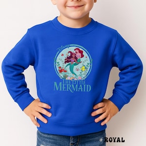 May include: Royal blue sweatshirt featuring a circular graphic of Ariel and other characters from Disney's The Little Mermaid. The graphic includes the Disney logo and the words "THE LITTLE MERMAID" in teal. The sweatshirt is a solid royal blue color.