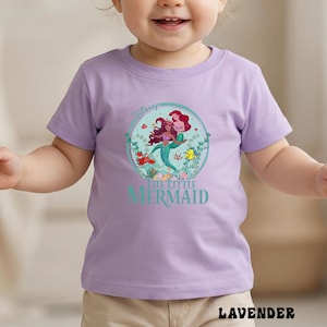 May include: A lavender short-sleeved t-shirt featuring a graphic of Ariel from Disney's The Little Mermaid. The design includes Ariel, Flounder, and Sebastian, surrounded by aquatic plants. The text "THE LITTLE MERMAID" is below the graphic.