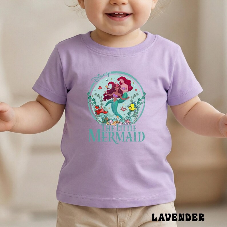 May include: A lavender short-sleeved t-shirt featuring a graphic of Ariel from Disney's The Little Mermaid. The design includes Ariel, Flounder, and Sebastian, surrounded by aquatic plants. The text "THE LITTLE MERMAID" is below the graphic.