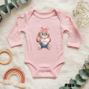 Peut inclure: Un body de b&eacute;b&eacute; rose clair &agrave; manches longues. Le devant pr&eacute;sente un lapin de dessin anim&eacute; tenant un c&oelig;ur avec le mot "love" dessus. Le lapin porte un n&oelig;ud rose et du denim bleu. Le body est en mati&egrave;re douce.