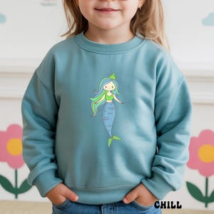 Puede incluir: Sudadera de color turquesa con una ilustraci&oacute;n de sirena de dibujos animados. La sirena tiene pelo azul y verde, un top verde y una cola azul. La palabra "CHILL" est&aacute; impresa en negro en la parte inferior derecha.