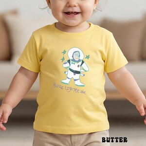 May include: A yellow t-shirt featuring a cartoon illustration of Buzz Lightyear in a spacesuit, with the text "Buzz Lightyear" in a playful font. The shirt is made of a soft material and is suitable for children.