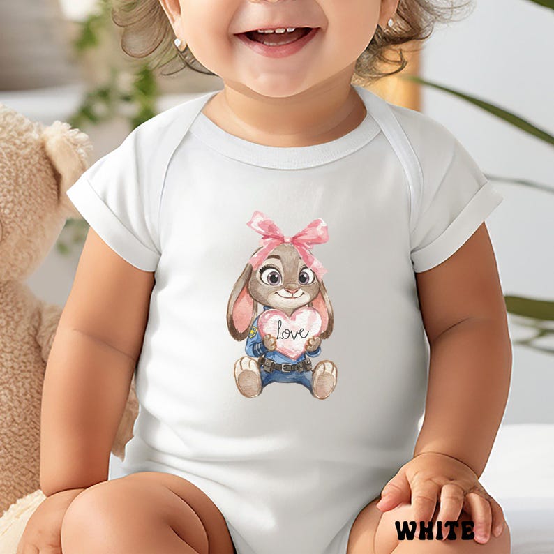 Peut inclure: Body b&eacute;b&eacute; blanc avec une illustration &agrave; l'aquarelle d'un lapin tenant un c&oelig;ur avec le mot "Love". Le lapin porte un n&oelig;ud rose et un uniforme bleu. Le mot "WHITE" est imprim&eacute; en bas.