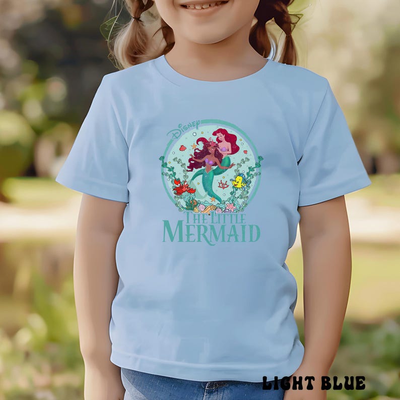 May include: Light blue t-shirt featuring a graphic from Disney's The Little Mermaid. The design includes Ariel and other characters in a circular frame with the text "The Little Mermaid". The shirt is light blue.