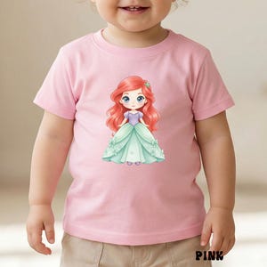 Puede incluir: Una camiseta de manga corta de color rosa claro con una ilustraci&oacute;n central de un personaje de dibujos animados con pelo largo rojo, un corpi&ntilde;o morado y un vestido de gala verde claro. La palabra PINK aparece en letras negras abajo.