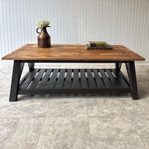 May include: A rectangular coffee table with a stained wood top and a black slatted shelf. A bronze vase with white flowers and two books are on the table. The table is approximately 122 cm long and 61 cm wide.