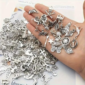 Puede incluir: Una colección de abalorios plateados de varias formas, incluyendo una calavera y huesos cruzados, una mariposa, una estrella y un ancla. Los abalorios se sostienen en una mano, con una pila más grande de abalorios en el fondo. Para hacer joyas.
