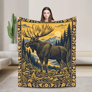 May include: A rectangular throw blanket featuring a large moose standing in a forest landscape with mountains and a full moon in the background. The design is rendered in shades of yellow and dark blue, framed by a decorative floral border pattern.
