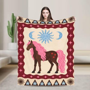 Könnte beinhalten: Eine cremefarbene Wohndecke mit einem braunen Pferd, das eine lange rosa Mähne und einen rosa Schweif hat. Das Design zeigt eine blaue Sonne und zwei Mondsicheln, umrahmt von einem dunkelroten Rand mit rosa Sternen und blauen Blumen.