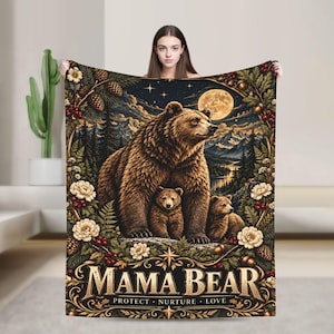 May include: A rectangular throw blanket featuring a large brown bear with two cubs in a forest landscape under a full moon. The design includes pine cones, berries, and white flowers. The text MAMA BEAR PROTECT NURTURE LOVE is at the bottom.