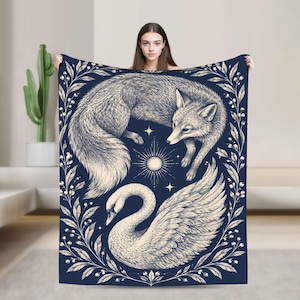 Peut inclure: Un plaid rectangulaire sur fond bleu foncé avec une illustration crème représentant un renard enroulé au-dessus d'un cygne. Le motif est encadré par une bordure botanique de feuilles et de baies, avec une icône de soleil entre les animaux.