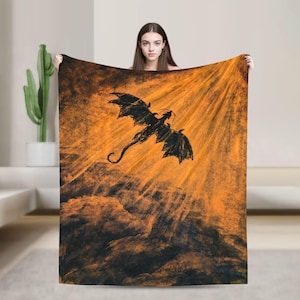 May include: A rectangular throw blanket featuring a black silhouette of a flying dragon centered against a vibrant orange and dark brown background with radiating light beams emanating from the upper right corner across the textured surface.