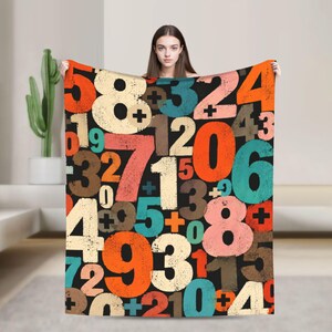 May include: A rectangular throw blanket featuring a black background covered in a repeating, distressed-style pattern of large numbers and plus signs in various colors including orange, teal, cream, brown, and pink across the entire surface.