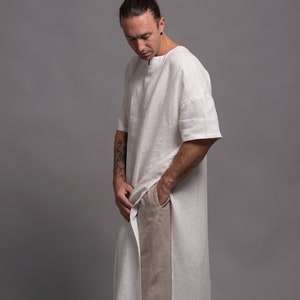 May include: A white linen tunic with a long, flowing design. The tunic has a round neckline and short sleeves. The model is wearing the tunic with beige linen trousers.