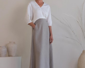 May include: A white V-neck top with three-quarter length sleeves paired with a long, high-waisted grey skirt. The skirt features a flared silhouette and side pockets. The ensemble is composed of a solid-coloured, textured fabric.