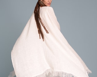 May include: A white linen poncho with a fringed edge. The poncho is worn over a white linen dress. The model has long brown dreadlocks.
