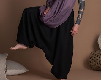 Può includere: Una persona indossa pantaloni neri con una vestibilità ampia e fluida e una sciarpa viola drappeggiata intorno al collo. La persona è in piedi con una gamba sollevata e le braccia lungo i fianchi.