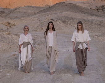May include: Three people wearing loose-fitting, white linen clothing with brown belts walk in a desert landscape. The clothing has a draped, flowing style.