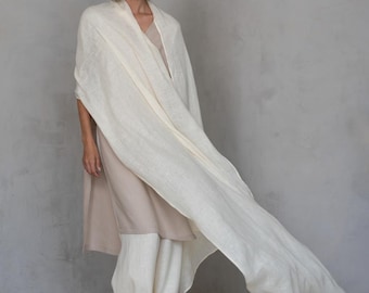 Puede incluir: Una mujer con un chal de lino color crema, un vestido beige y pantalones blancos. El chal está drapeado sobre los hombros y ondea al viento. El atuendo es sencillo y elegante, con una estética minimalista.