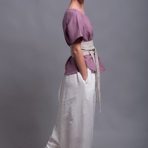 May include: A woman wearing a loose-fitting, mauve linen top with a wide, beige linen belt. She is also wearing white linen pants with a gathered elastic waistband. The pants are wide-legged and flowy. The model is standing on a gray background. The text "shantimama URBAN FOLK WEAR" is visible at the top of the image.