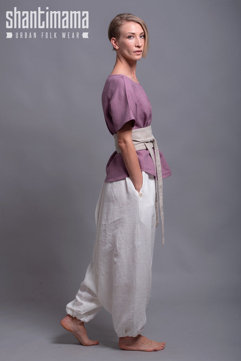 May include: A woman wearing a loose-fitting, mauve linen top with a wide, beige linen belt. She is also wearing white linen pants with a gathered elastic waistband. The pants are wide-legged and flowy. The model is standing on a gray background. The text "shantimama URBAN FOLK WEAR" is visible at the top of the image.