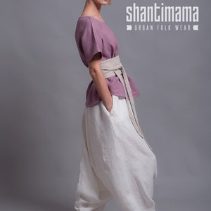 May include: A woman wearing a loose-fitting, dusty rose linen top with a wide, beige linen belt. She is also wearing white linen pants with a gathered detail at the ankles. The pants have a large, loose fit. The image has the text "shantimama URBAN FOLK WEAR" at the top.
