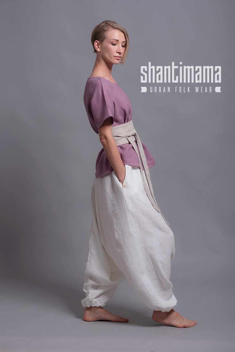 May include: A woman wearing a loose-fitting, dusty rose linen top with a wide, beige linen belt. She is also wearing white linen pants with a gathered detail at the ankles. The pants have a large, loose fit. The image has the text "shantimama URBAN FOLK WEAR" at the top.