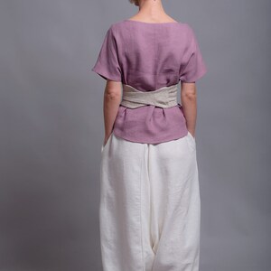 May include: A woman wearing a mauve linen top with a beige linen belt and white linen pants. The top has short sleeves and a scoop neck. The pants are loose fitting and have a wide leg. The woman is standing with her hands in her pockets. The text "shantimama URBAN FOLK WEAR" is visible at the top of the image.