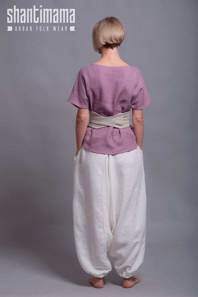May include: A woman wearing a mauve linen top with a beige linen belt and white linen pants. The top has short sleeves and a scoop neck. The pants are loose fitting and have a wide leg. The woman is standing with her hands in her pockets. The text "shantimama URBAN FOLK WEAR" is visible at the top of the image.