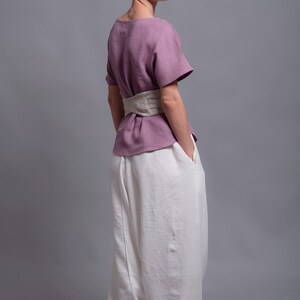 May include: A woman wearing a mauve linen top with a beige linen belt and white linen pants. The top has a scoop neck and short sleeves. The pants are loose fitting and have a wide leg. The woman is standing with her back to the camera and her hands in her pockets. The text "shantimama URBAN FOLK WEAR" is visible at the top of the image.