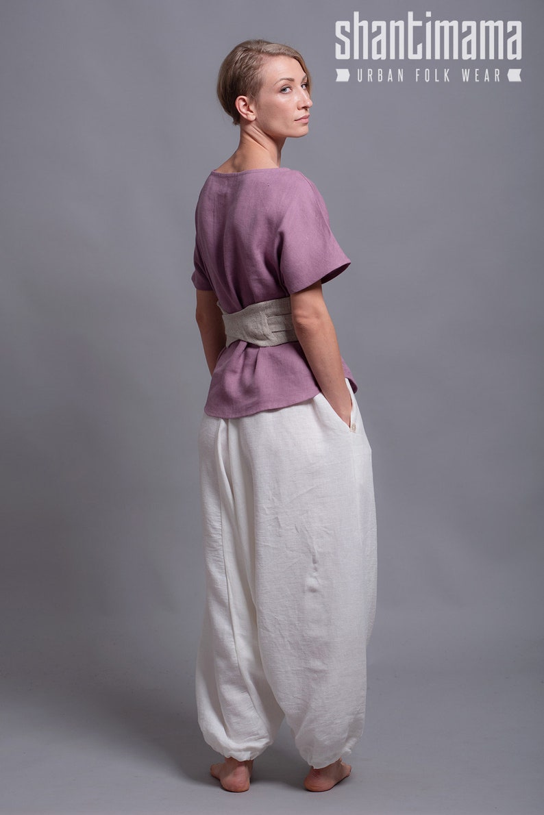 May include: A woman wearing a mauve linen top with a beige linen belt and white linen pants. The top has a scoop neck and short sleeves. The pants are loose fitting and have a wide leg. The woman is standing with her back to the camera and her hands in her pockets. The text "shantimama URBAN FOLK WEAR" is visible at the top of the image.