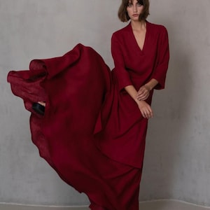 May include: A flowing, deep red dress with a V-neck and three-quarter sleeves. The dress is made of a lightweight fabric that drapes elegantly. The model is wearing black sandals. The background is a neutral gray.