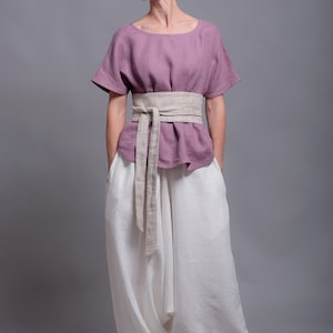 May include: A woman wearing a mauve linen top with a beige linen belt and white linen pants. The pants are loose fitting and have a wide leg. The woman is standing on a gray background. The text "shantimama URBAN FOLK WEAR" is visible in the top right corner of the image.
