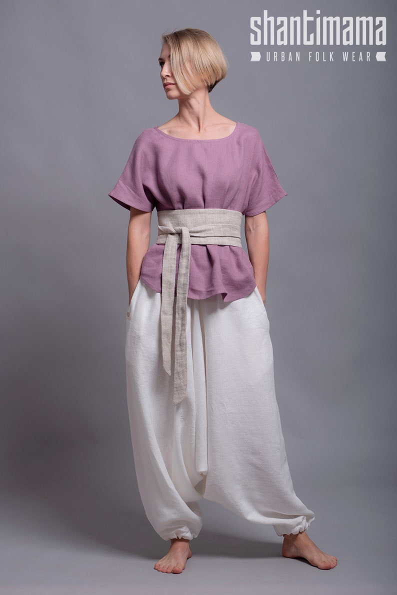 May include: A woman wearing a mauve linen top with a beige linen belt and white linen pants. The pants are loose fitting and have a wide leg. The woman is standing on a gray background. The text "shantimama URBAN FOLK WEAR" is visible in the top right corner of the image.