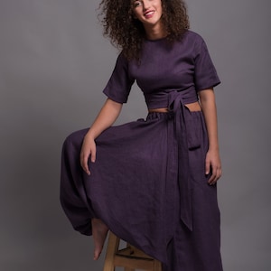May include: A woman wearing a purple linen crop top and matching wide-leg pants. The top has a tie at the waist and the pants have a loose, flowing fit. She is sitting on a wooden stool.
