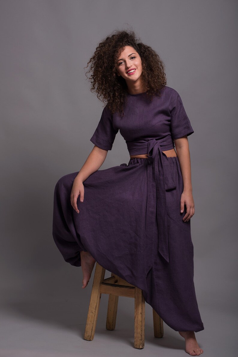 May include: A woman wearing a purple linen crop top and matching wide-leg pants. The top has a tie at the waist and the pants have a loose, flowing fit. She is sitting on a wooden stool.