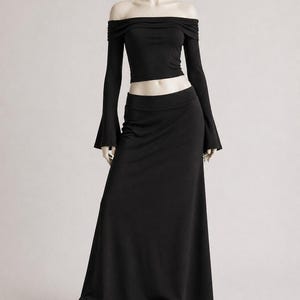 May include: A black off-the-shoulder top with long sleeves and a matching floor-length skirt. The top has a fitted silhouette, and the skirt flows to the ground. The outfit is paired with black and white sneakers.