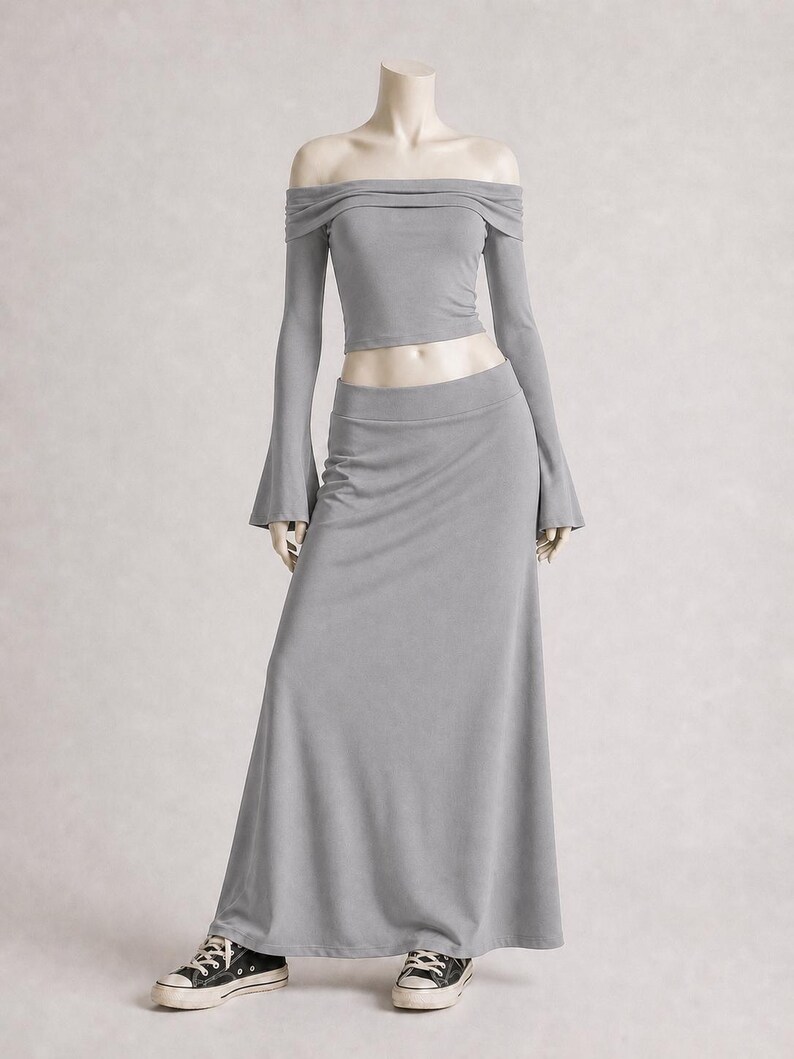 May include: A gray two-piece outfit on a mannequin. The top is an off-the-shoulder crop top with long sleeves. The bottom is a long, flowing skirt. The mannequin is wearing black and white sneakers.