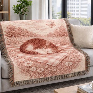 May include: A woven throw blanket with a pastoral scene in shades of red and cream, draped over a gray sofa. The blanket features a sleeping cat, a butterfly, and a floral border. The throw has a fringed edge.