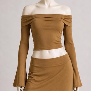 May include: A two-piece set in a warm brown hue. The off-the-shoulder top features long sleeves and a gathered neckline. The matching skirt has a high waist. The fabric appears to be a soft, suede-like material.