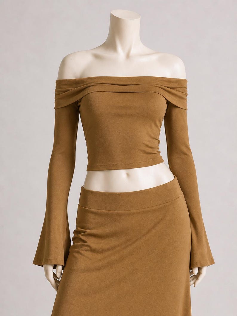May include: A two-piece set in a warm brown hue. The off-the-shoulder top features long sleeves and a gathered neckline. The matching skirt has a high waist. The fabric appears to be a soft, suede-like material.