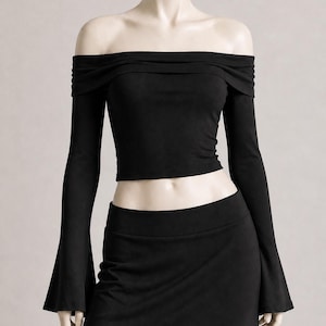 May include: A black off-the-shoulder crop top with long sleeves and a matching black skirt. The top has a gathered neckline and the sleeves flare out at the wrists. The skirt is a simple, fitted design.