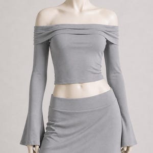 May include: A gray off-the-shoulder top with long sleeves and a matching skirt. The top has a draped neckline and the sleeves flare out at the wrists. The skirt is a high-waisted, fitted design. The outfit is displayed on a mannequin.
