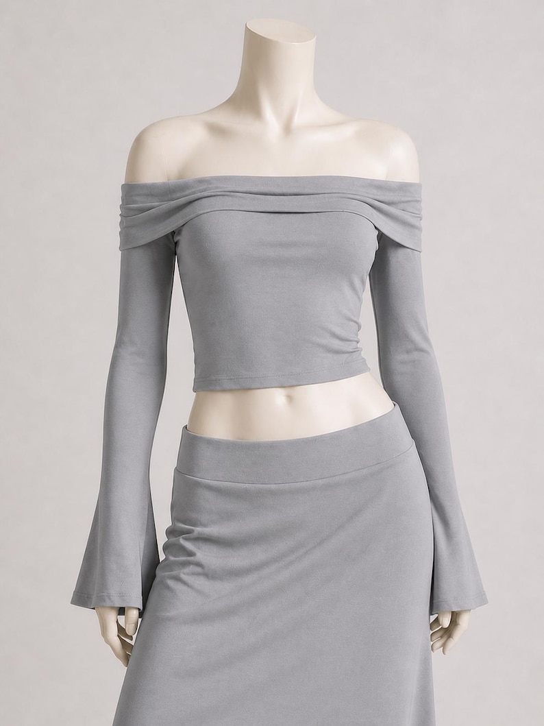 May include: A gray off-the-shoulder top with long sleeves and a matching skirt. The top has a draped neckline and the sleeves flare out at the wrists. The skirt is a high-waisted, fitted design. The outfit is displayed on a mannequin.