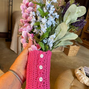 May include: A bouquet of artificial flowers in shades of pink, blue, and purple, held in a bright pink crocheted sleeve with white buttons. The sleeve is designed to hold a vase or container.