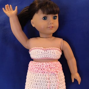May include: A doll wearing a handmade crochet outfit. The outfit includes a bandeau top and a skirt, both in white with pink accents. The skirt has a pink tie. The doll has brown hair and blue eyes.