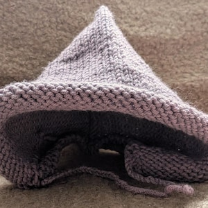 May include: A lavender-coloured, hand-knitted hat with a pointed top and a wide brim. The hat has a textured knit pattern and a drawstring closure. The interior of the hat is a darker shade of purple. This cosy hat is suitable for costume or everyday wear.