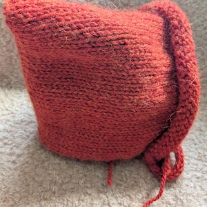 May include: A rust-coloured, hand-knitted hat with a pointed top and a rolled brim. It has a long, braided cord for tying. The textured knitwear is made of a soft, warm material.