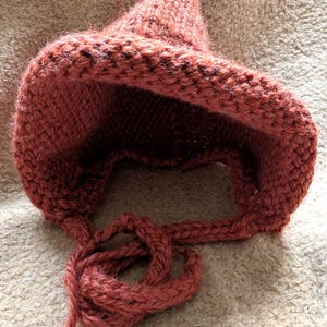 May include: A hand-knitted bonnet in a muted terracotta orange hue, featuring a textured garter stitch pattern, a pointed crown, and two thick braided ties extending from the base to secure the head covering under the chin.